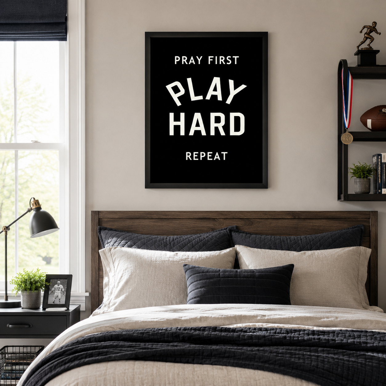Black framed vertical Christian athlete wall art featuring Pray First Play Hard Repeat in bold cream varsity typography displayed as a 20x26 statement piece above a dark wood headboard in a teen boy's sports bedroom with football trophy medal and navy bedding