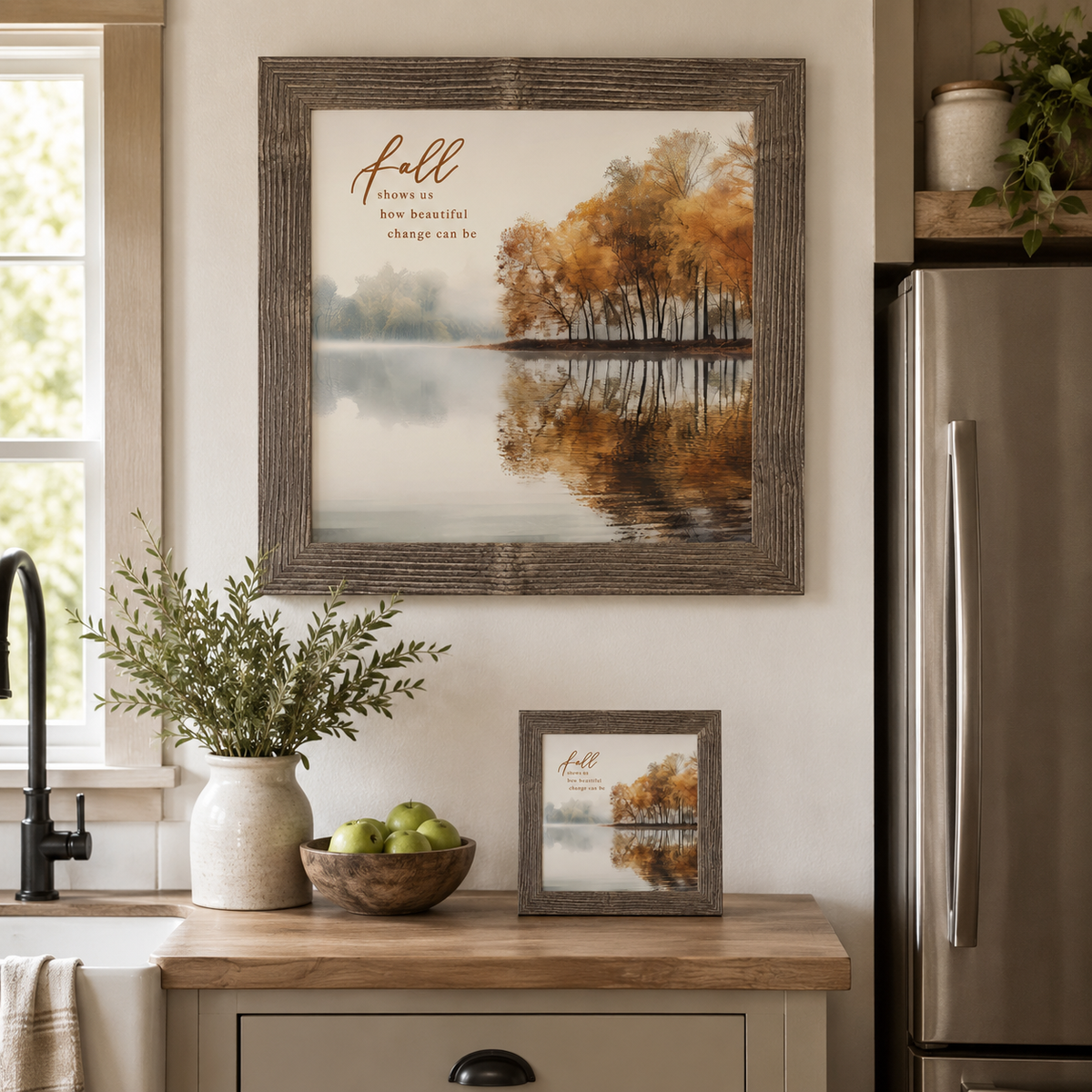 Natural brown framed square fall wall art displayed in a farmhouse kitchen above the counter with a matching smaller piece on an easel beside a ceramic vase and bowl of green apples, featuring a misty autumn lake with golden amber trees and the words "Fall Shows Us How Beautiful Change Can Be"