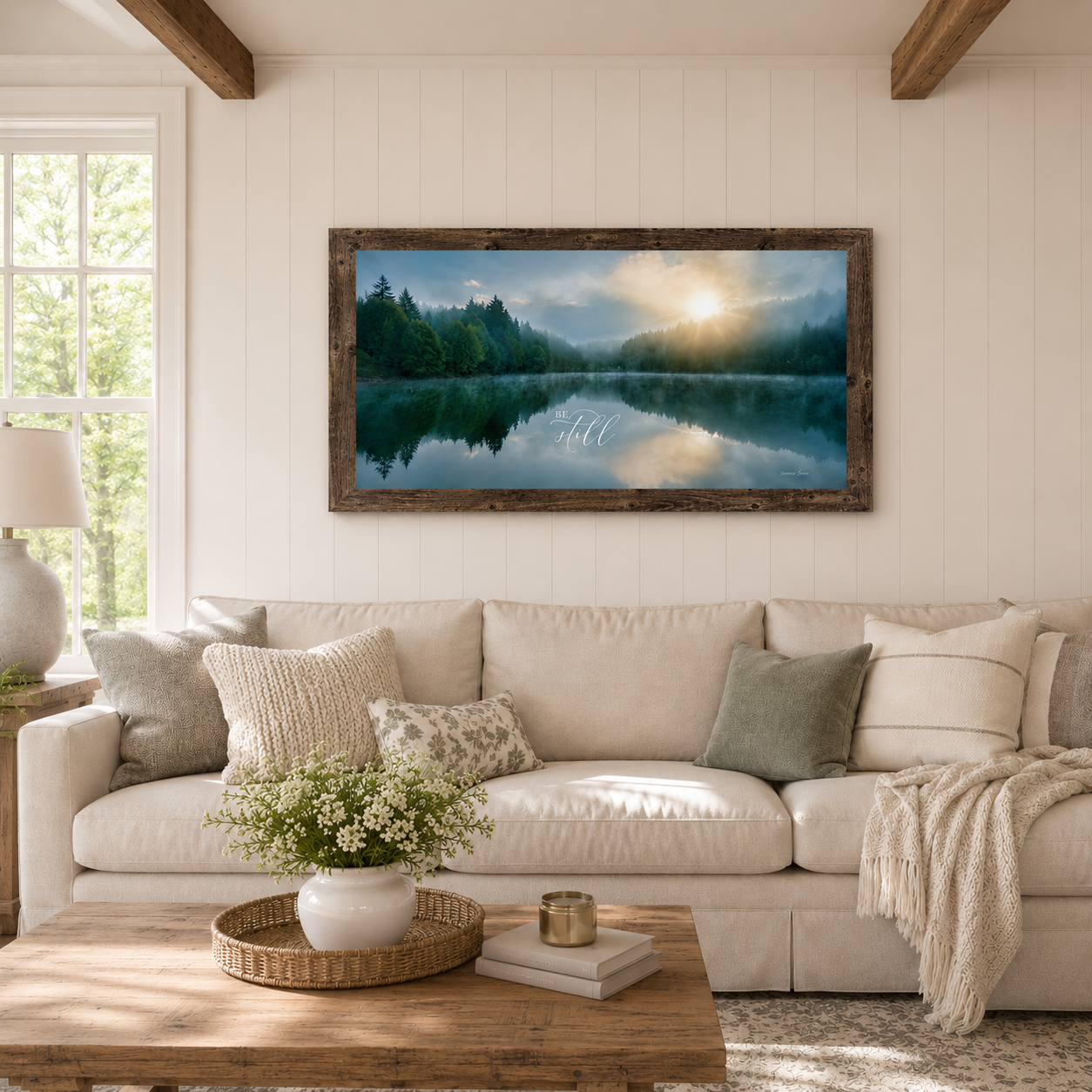 Natural brown farmtable framed Be Still wide landscape wall art SB765 displayed large above cream linen sofa in bright farmhouse living room with shiplap walls wood beam ceiling and white floral centerpiece — misty lake sunrise with pine forest reflection and golden sun breaking through clouds with elegant Be Still script typography