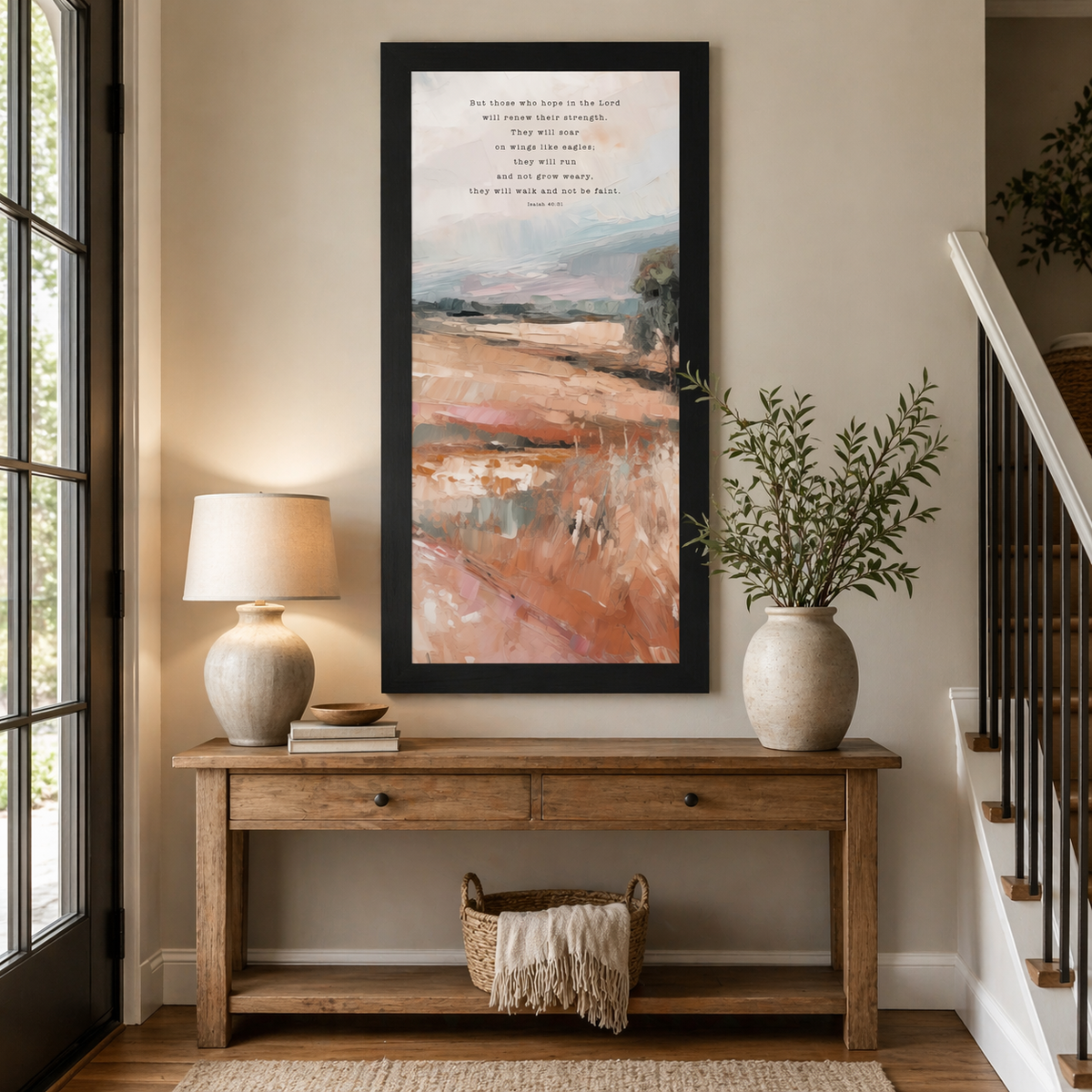 Black framed tall portrait Christian wall art displayed at large scale in a warm neutral entryway with wooden console table beside staircase showing Isaiah 40:31 soar on wings like eagles scripture in typewriter typography over soft peach blush terracotta painterly landscape