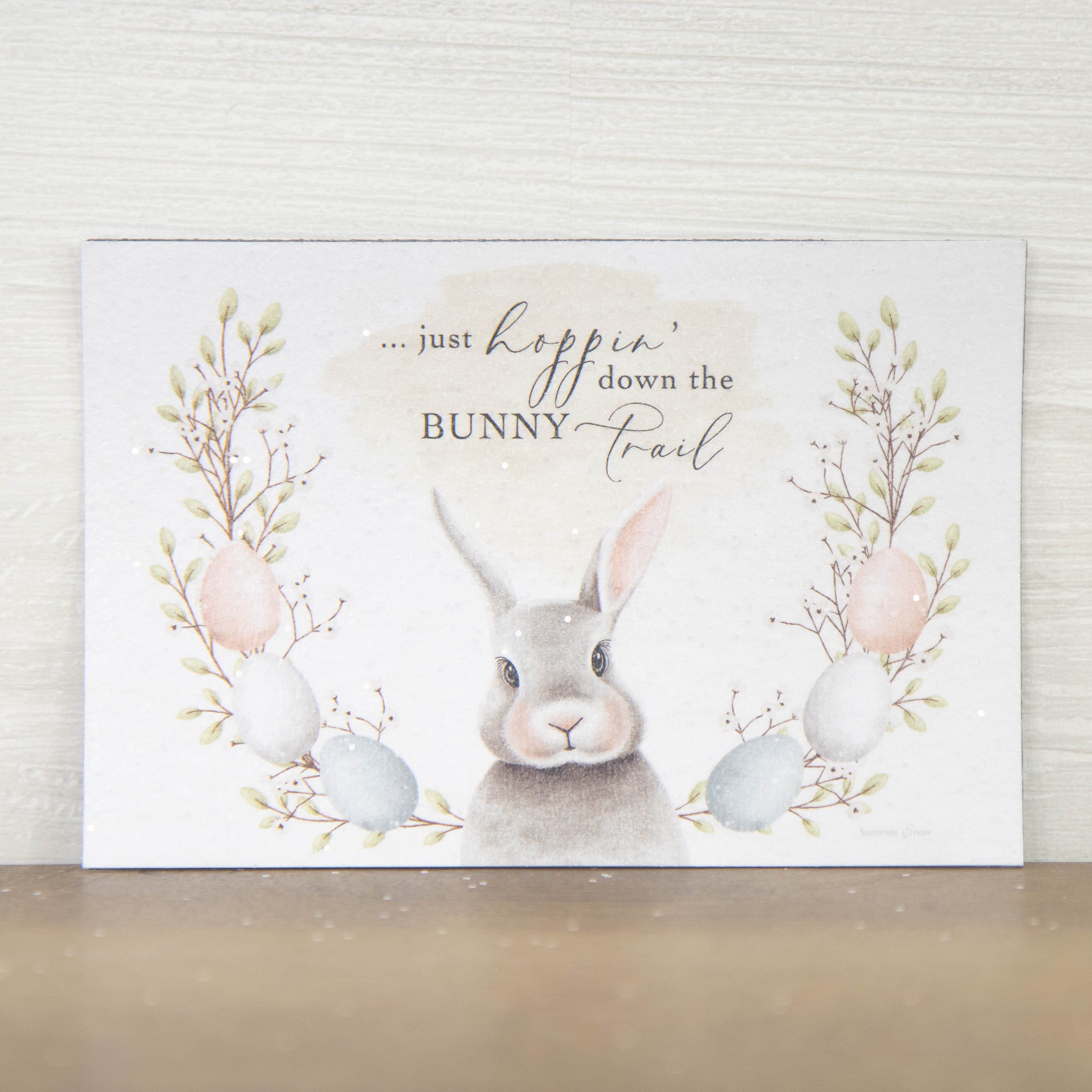 Cute Easter magnet featuring gray bunny with egg and floral wreath, Just hoppin’ down the bunny trail quote on neutral background with subtle glitter finish
