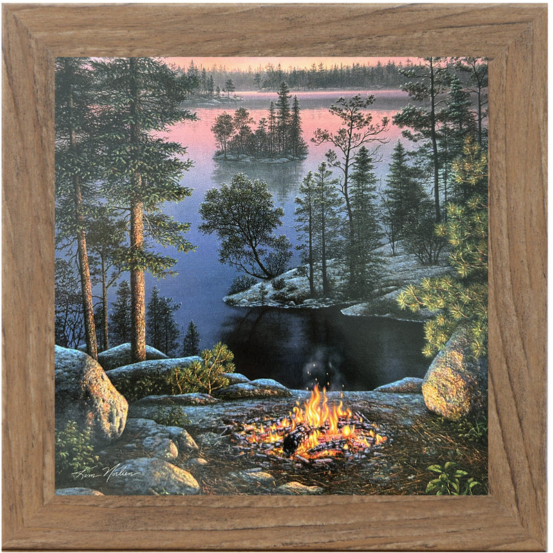 Framed 8x8 wall art showing a lakeside campfire at sunset with calm water, pine trees, and distant hills in soft golden tones. Art by Kim Norlien.